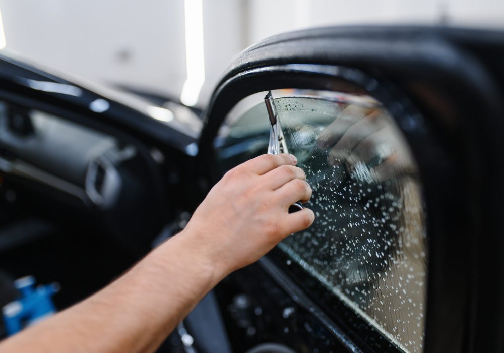 Male worker with squeegee wipes car tinting, tuning service. Mechanic applying vinyl tint on vehicle window in garage, tinted automobile glass